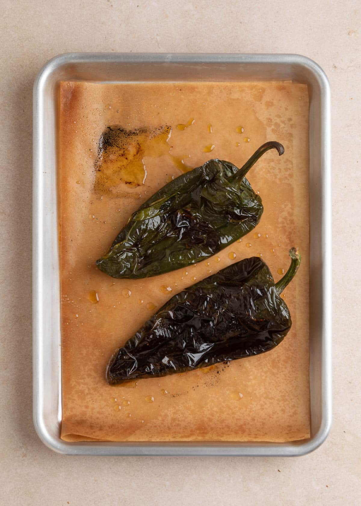 Two whole poblano peppers with dark, wrinkled, blistered skins and charred patches rest on a parchment-lined metal baking sheet with scattered droplets of olive oil.