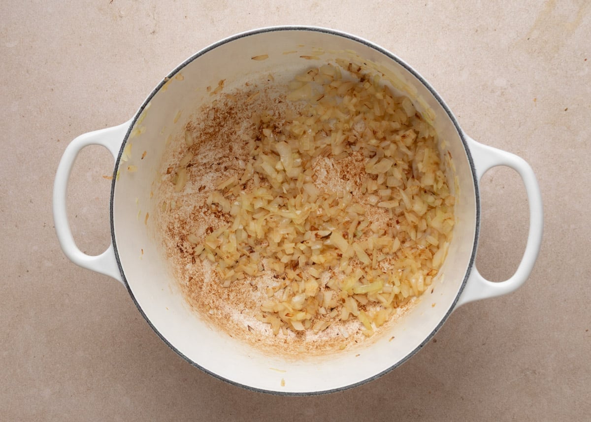 Diced onions softened until translucent with lightly golden edges in a white enameled Dutch oven, with browned bits visible on the bottom of the pot.