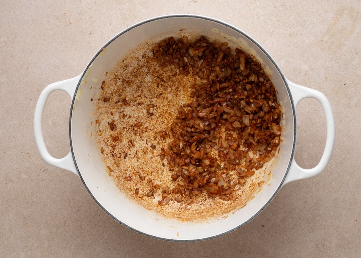 Saut&eacute;ed diced onions stirred together with spices, coating the onions in a reddish-brown seasoning mixture in a white enameled Dutch oven.
