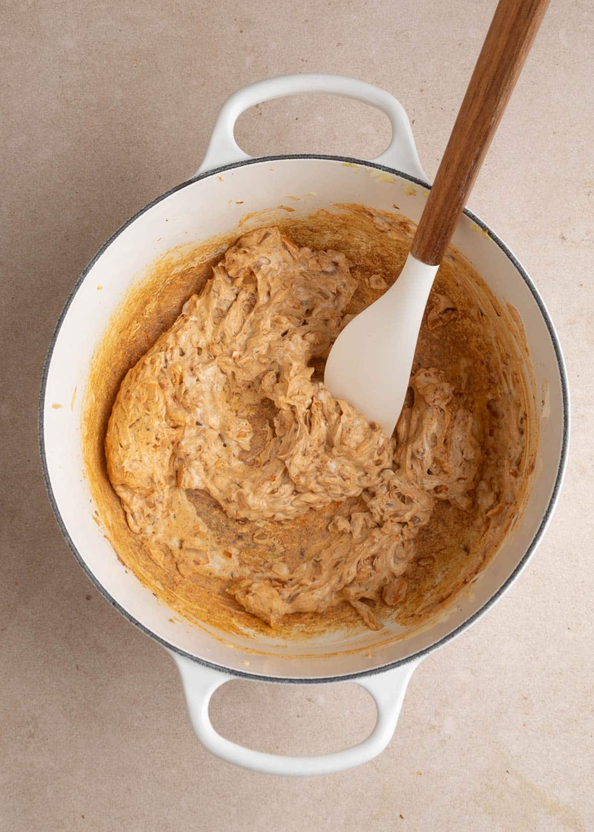 Thick, creamy, orange-tinted mixture of softened cream cheese, saut&eacute;ed onions, and spices being stirred with a white silicone spatula in a white enameled Dutch oven.